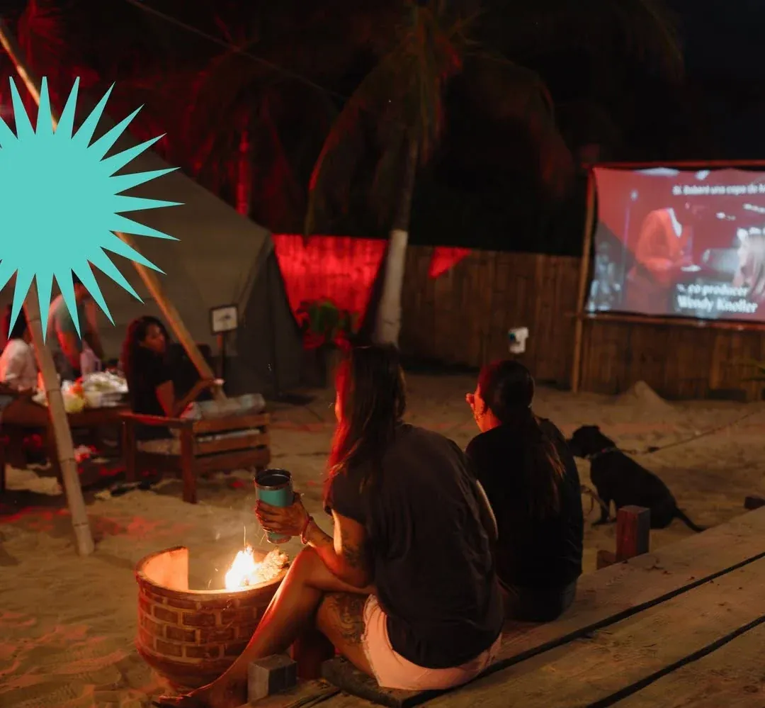 Guests around the bonfire at night, with the outdoor cinema screen glowing behind them