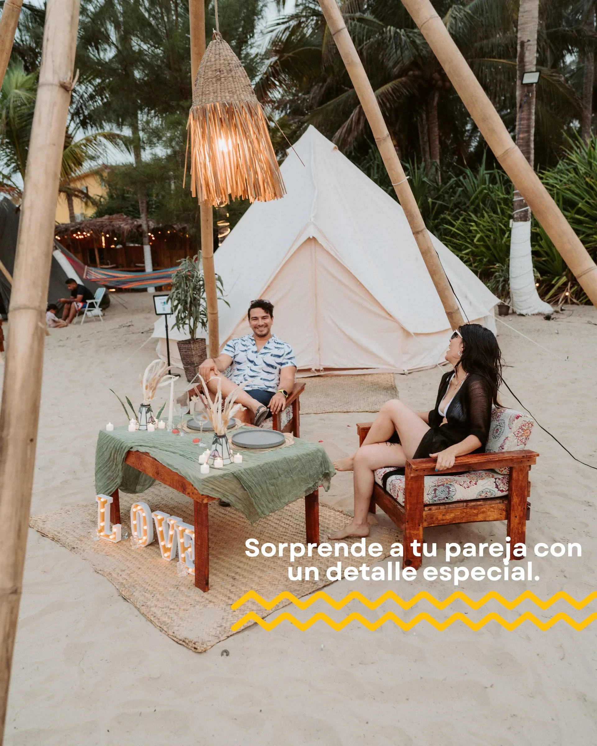 A couple talking in front of their tent, beside a LOVE letter setup on the sand